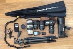 Photography equipment on a wooden floor, including two cameras, multiple lenses, a tripod, a light stand, an umbrella, and various camera accessories.