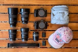 Camera gear and accessories arranged on a wooden surface, including two cameras, five lenses, a strap, two batteries, a gray pouch, and a multicolored hat.