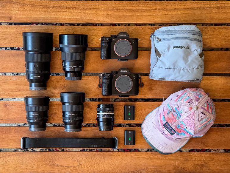 Camera gear and accessories arranged on a wooden surface, including two cameras, five lenses, a strap, two batteries, a gray pouch, and a multicolored hat.