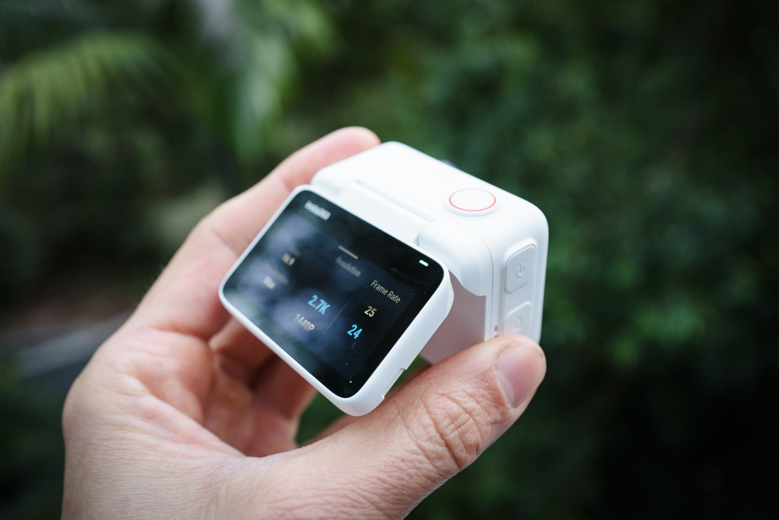 A person holds a compact digital device with a small screen displaying data. The device is white with buttons on the side and a red ring on top. The background is blurred greenery.