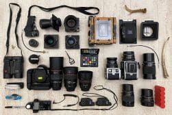 A collection of photography equipment, including cameras, lenses, binoculars, a wooden camera, and various accessories, arranged neatly on a light wooden surface.