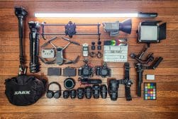 Various filmmaking equipment neatly arranged on a wooden floor, including a drone, cameras, lenses, microphones, a light panel, a slate clapper, and other accessories.
