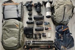 Photography equipment laid out on a wooden floor, including several cameras, lenses, backpacks, and tripods.
