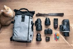 Camera equipment and accessories, including several lenses, a grey backpack, a video camera, and colorful balls, laid out on the floor. A cat lies nearby.