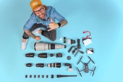 A person with glasses, a beard, and a yellow hat sits on a blue background, giving a thumbs-up next to an array of camera gear, including lenses, a drone, and other photography equipment.