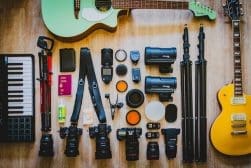Assorted photography and music equipment, including cameras, lenses, tripods, microphones, a keyboard, a green guitar, and a yellow guitar, organized neatly on a wooden floor.