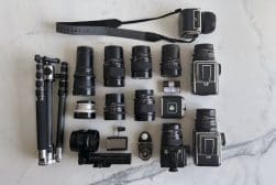 Camera equipment including multiple lenses, two camera bodies, a strap labeled "Hasselblad", three tripods, and various accessories arranged on a white surface.
