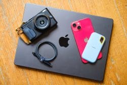 A black laptop with the Apple logo, a black camera, a red smartphone, a blue and white wallet-sized device, and a black USB cable are arranged on a wooden surface.