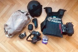 A collection of press equipment on a wooden floor. Items include a helmet, tactical vest labeled "PRESS," camera, lenses, microphone, memory cards, and a beige bag.