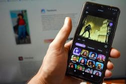 A person holds a smartphone open to an app displaying various photo filter options. In the background, a computer screen is seen with another image and text.