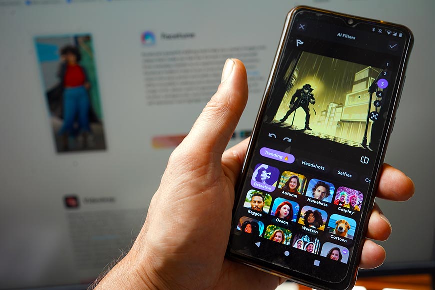A person holds a smartphone open to an app displaying various photo filter options. In the background, a computer screen is seen with another image and text.
