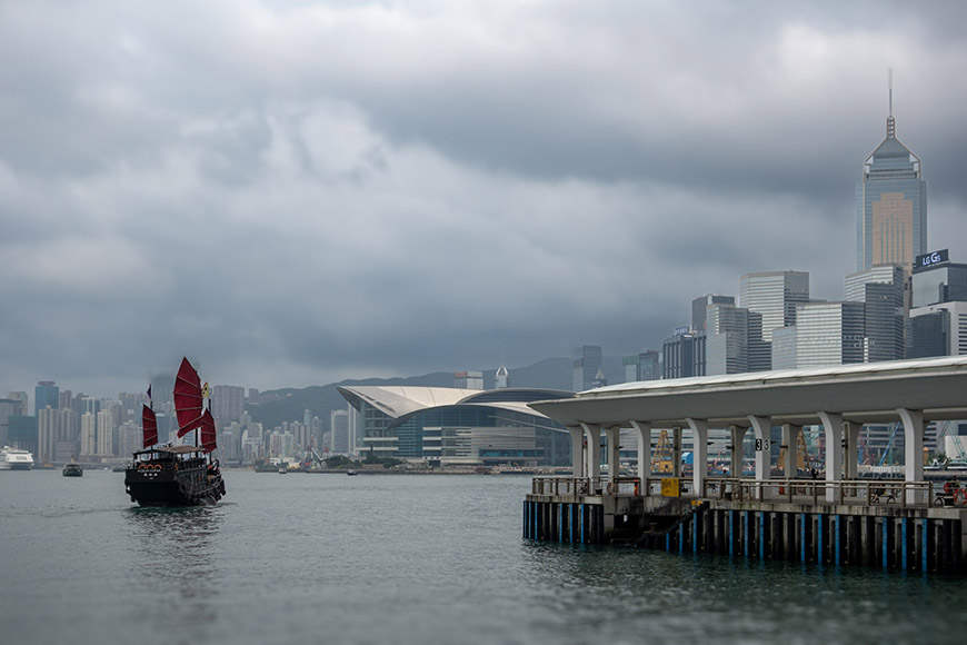 Traditional junk boat with red sails on water near a modern city skyline under cloudy skies. A pier is visible on the right.