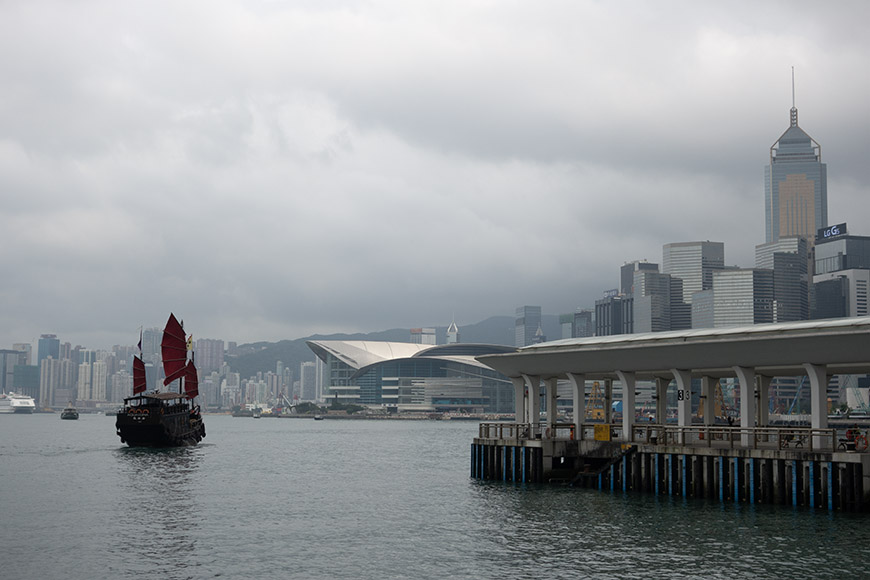 A traditional junk boat sails in Victoria Harbour near modern skyscrapers and a pier, under a cloudy sky in Hong Kong.
