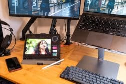A workspace setup with two monitors, a tablet displaying a photo-editing app, a laptop on a stand, a keyboard, a stylus, headphones, and a smartphone on a wooden desk.