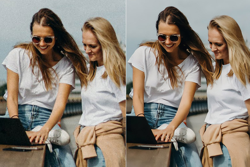 Two women sitting outdoors, looking at a laptop. One wears sunglasses and jeans, the other has a beige sweater tied around her waist. Both are smiling.