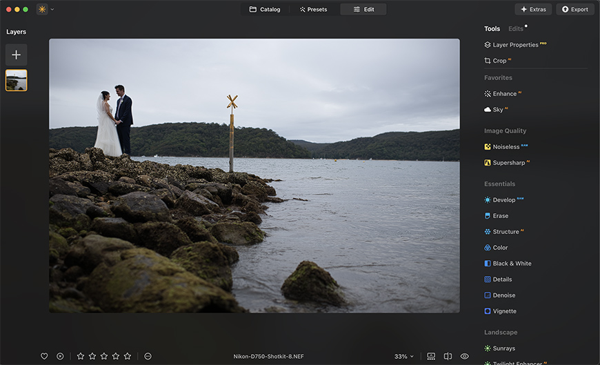 A couple in wedding attire stand on a rocky shore by a large body of water, with forested hills in the background. A cross-shaped sign is visible near the water's edge.