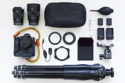 Flat lay of photography equipment including a camera, lenses, tripod, batteries, filters, cleaning supplies, and a carrying case, all arranged neatly on a white background.