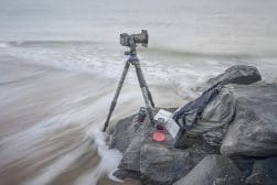 Camera on a tripod set up on rocks by the beach, with a backpack, notebook, and camera lenses nearby, surrounded by gentle waves.