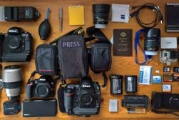 Camera equipment and accessories arranged on a wooden surface, including multiple cameras, lenses, batteries, memory cards, a press vest, and various tools for photography.