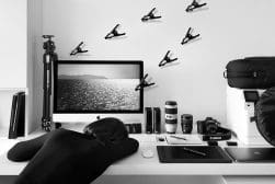 Person resting their head on a white desk with a computer showing a seascape. Various photography equipment, including lenses and a camera, are organized on the desk. Clamps are mounted on the wall.