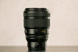 A Nikon Nikkor Z 135mm f/1.8 S lens is placed upright against a light pink backdrop.