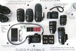 A collection of Fujifilm camera equipment, including cameras, lenses, film rolls, batteries, and accessories, neatly arranged on a flat surface next to a smartphone, glasses, and a coffee cup.