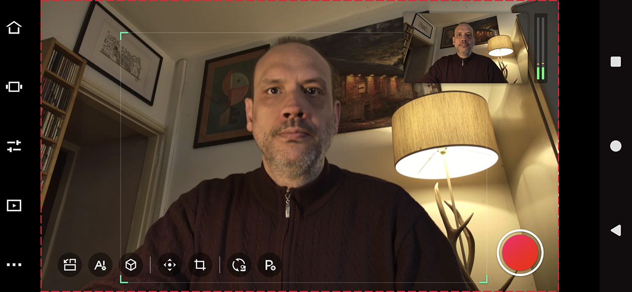 A man is sitting indoors in front of a lamp and framed art, with a smaller inset image of him in the top right corner. Various interface icons are visible on the screen.