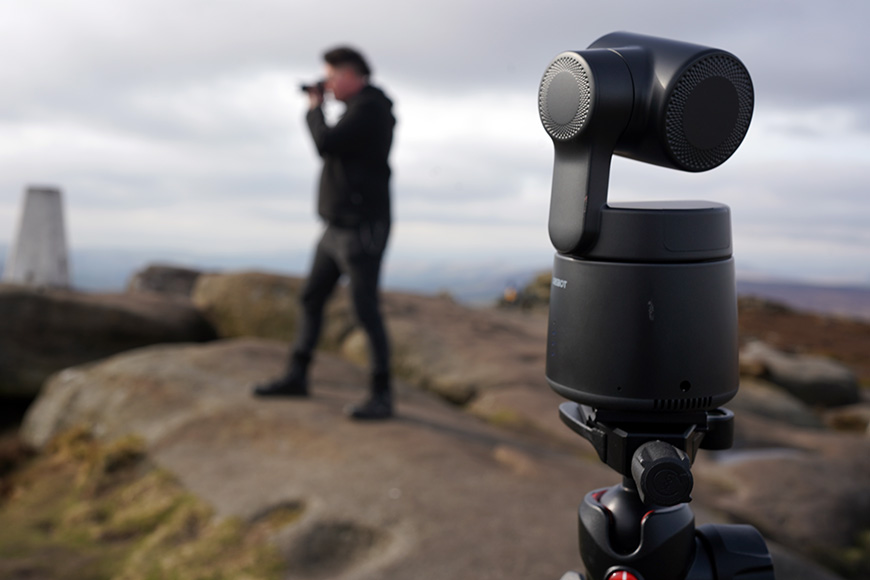 Close-up of a camera on a tripod with a person in the background taking a photo on a rocky landscape.
