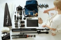 Overhead view of a photographer’s gear laid out, including cameras, lenses, tripods, lighting accessories, a tablet, and a backpack. A person is holding a mirror reflecting their face.