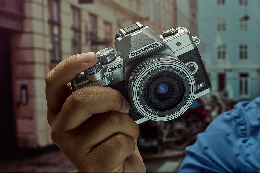 Close-up of a hand holding a silver Olympus OM-D camera against a blurred urban background.