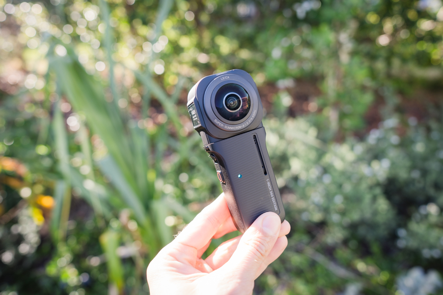 A person holding a small camera in front of bushes.