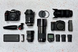 A collection of photography gear including cameras, lenses, a tripod, lens caps, a flash trigger, batteries, and a power bank arranged on a speckled surface.