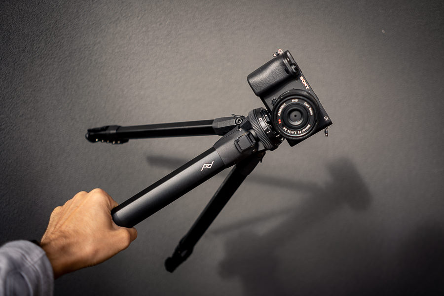 A person's hand holding a compact tripod with a camera mounted on it against a gray background.