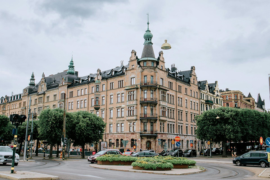 Historic European building with intricate architecture stands by a roundabout, surrounded by trees and traffic signs, under a cloudy sky.