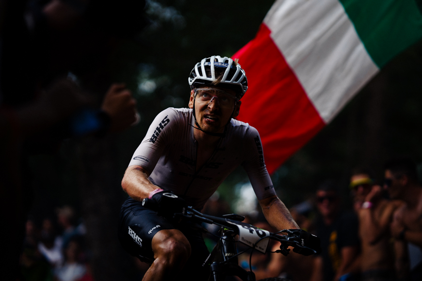 A cyclist in a helmet and cycling attire rides intensely during a race, with a large flag visible in the background.