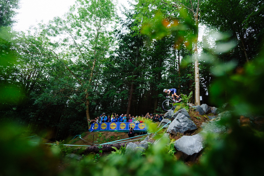 Cyclist rides downhill on a rocky trail in a forested area as spectators watch from behind a blue barrier.