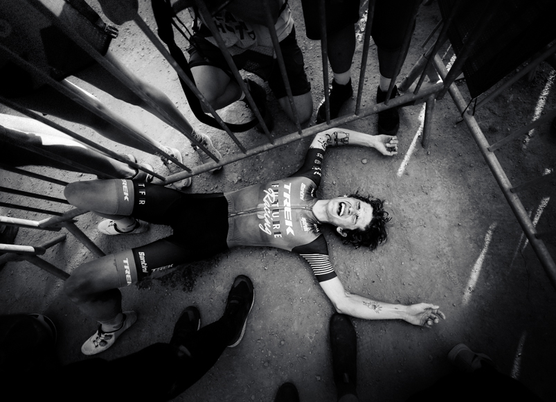 Cyclist wearing a team jersey lies on the ground near barriers, surrounded by people's legs.