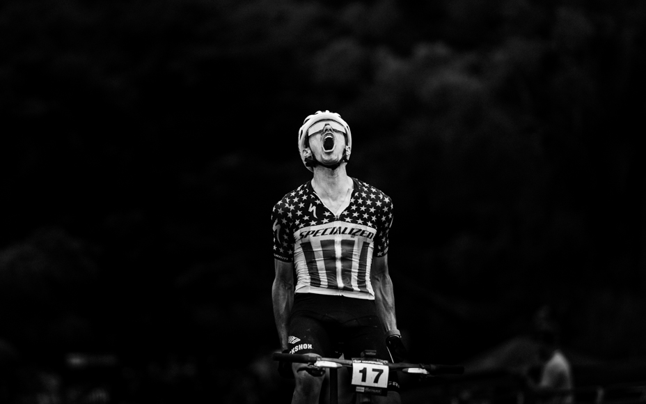 A cyclist wearing a star-spangled jersey and helmet raises their head, shouting, while riding a bike in a dark setting.