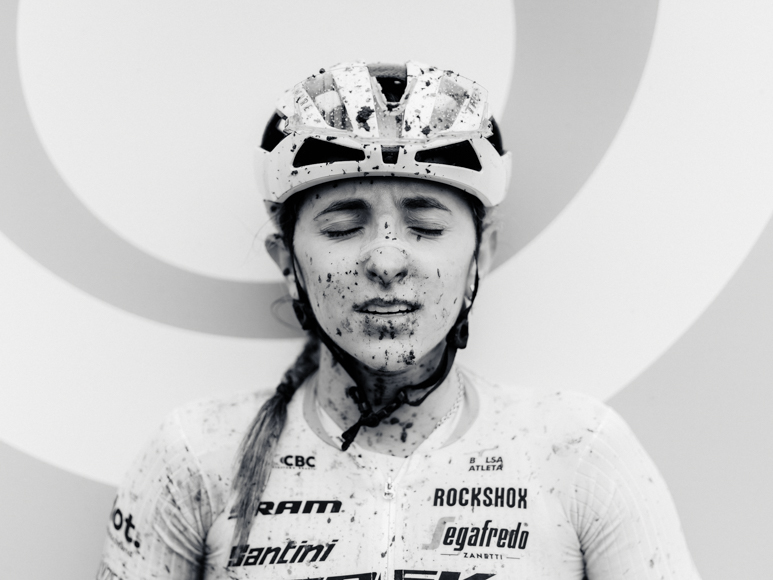Cyclist with closed eyes, wearing a helmet and jersey, covered in mud splatters against a circular patterned background.