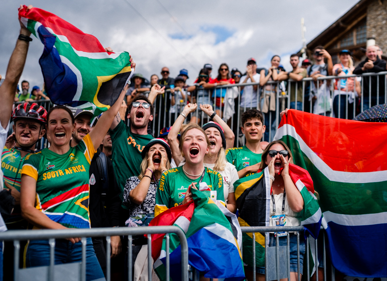 A cheering crowd waves South African flags and wears green jerseys behind a barrier at an outdoor event.