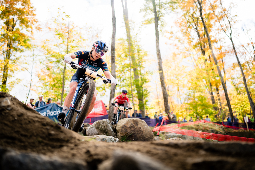 Cyclist navigating a rocky trail during a race in a sunlit forest, with another cyclist in the background.