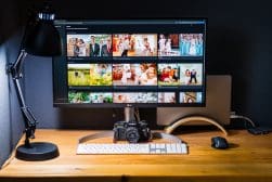 A computer monitor on a wooden desk displays a photo gallery featuring wedding images. A camera, keyboard, mouse, and desk lamp are also placed on the desk.