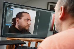 A person is viewing a close-up profile photo of a man being edited on a computer screen. The editing software interface is visible on the monitor.