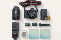 Camera equipment including a DSLR with lens, additional lens, film camera, flash, filter, batteries, and film roll, arranged on a white background.