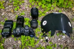 A digital camera with a lens, three additional lenses, a flash unit, and a carbon fiber bicycle helmet are placed on gravel with green plants around.