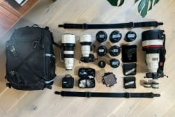 Photography equipment laid out on a wooden floor, including a backpack, multiple lenses, cameras, and tripods.
