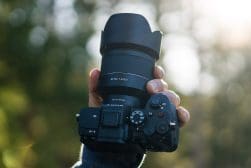 A hand holds a black DSLR camera with a large lens, surrounded by a softly blurred outdoor background.