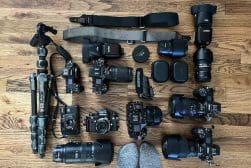 A collection of cameras, lenses, and accessories is arranged on a wooden floor, next to a pair of socked feet.