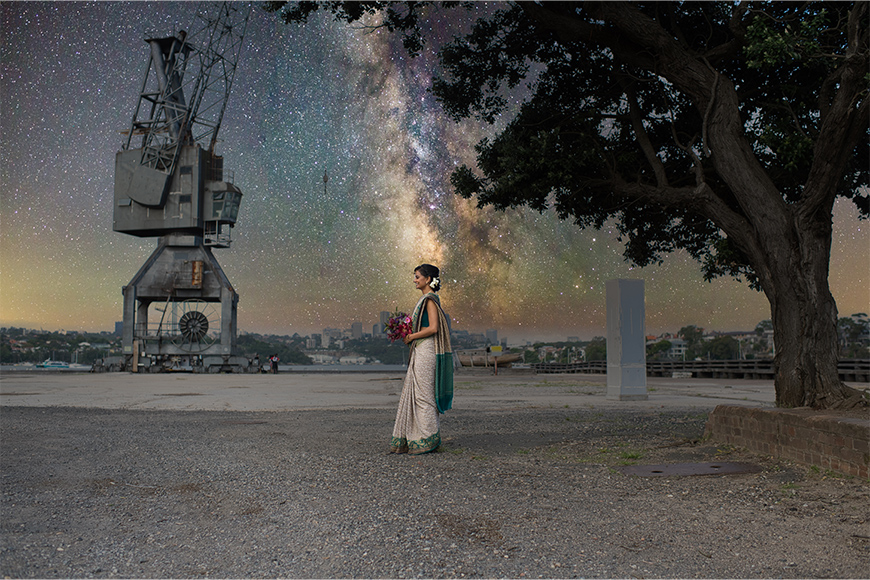 A woman in traditional attire stands by a tree on a waterfront, holding flowers. The night sky with stars and the Milky Way is visible above an industrial crane in the background.
