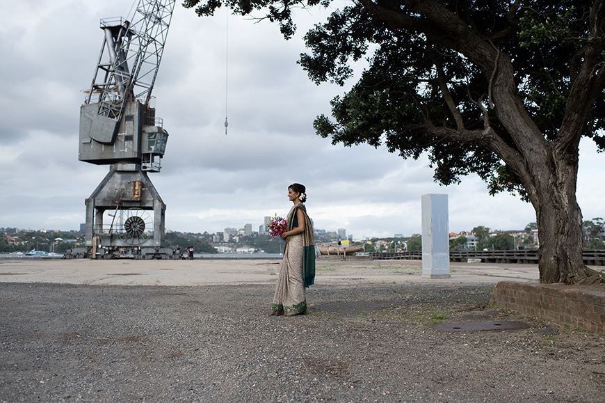 A person in traditional attire stands holding flowers near an industrial crane and a tree, with a cityscape in the background.